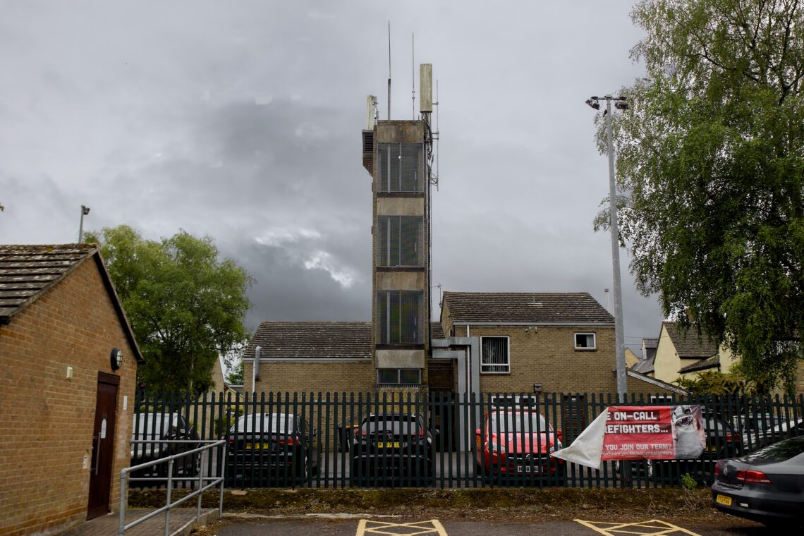 A three-storey beige brick fire station with a tall training tower stands behind a black metal fence. Several cars are parked outside. A red banner about on-call firefighters hangs on the fence. Trees and cloudy skies are visible in the background.
