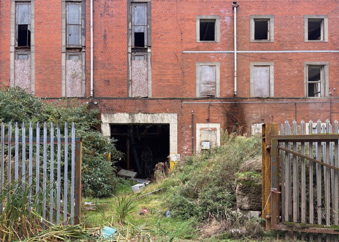 An abandoned red brick building with broken and boarded-up windows. Overgrown grass and bushes fill the foreground, along with metal gates, one of which is rusted and partly open. Rubbish and debris are visible near the building’s dark entrance.