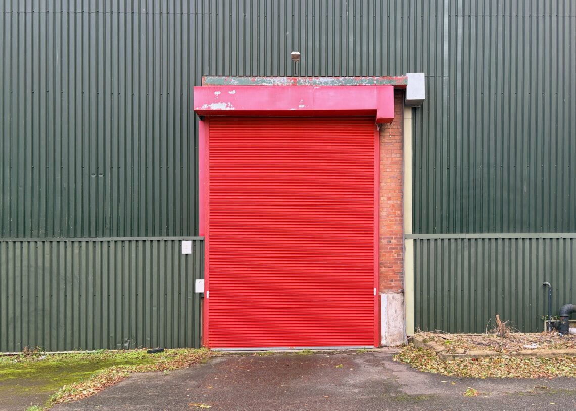 A bright red metal roller shutter door is closed on a green corrugated metal building. The frame above the door is also red, and the ground in front has some leaves and debris. The building shows minor signs of weathering.