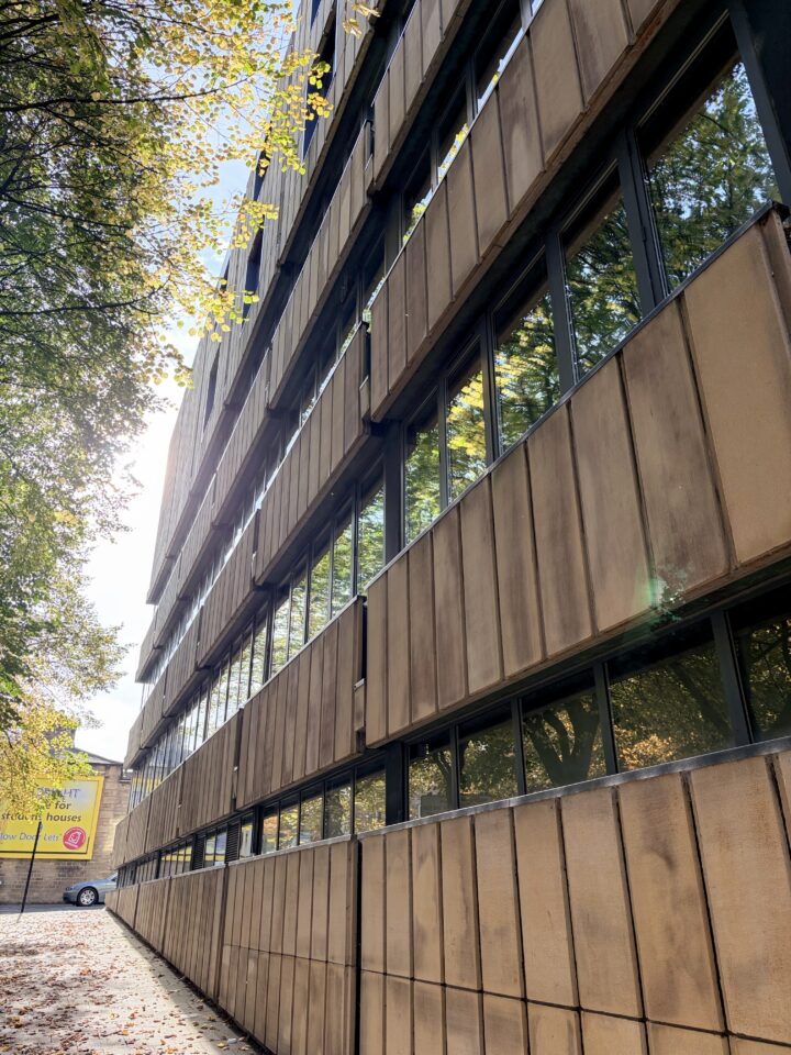 A low-angle view of a tan, modern building with large rectangular windows reflecting trees and sky. Sunlight filters through green-yellow leaves above. On the left, a parked white car and a yellow sign are visible in the distance.