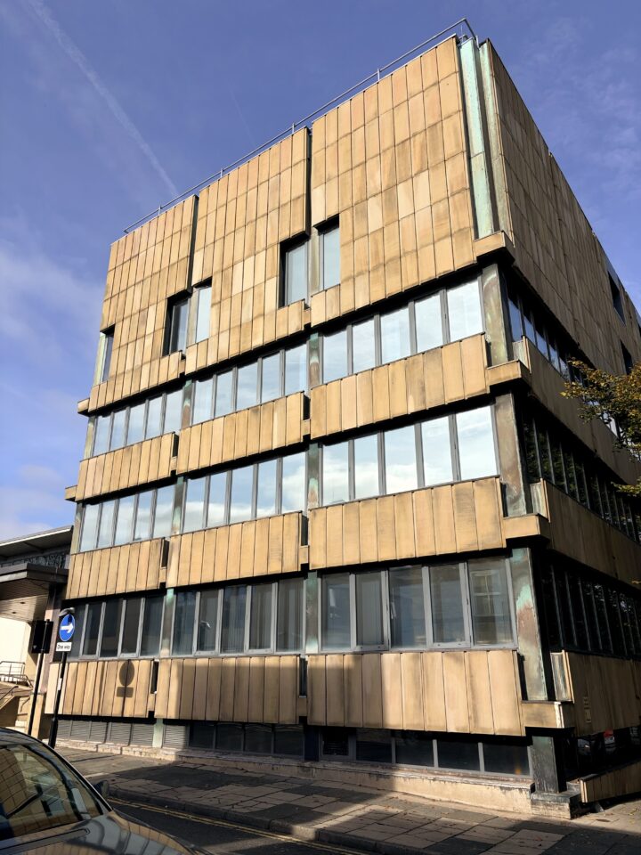 A four-storey building with a facade of yellowish-brown panels and many rectangular windows, set against a clear blue sky. There is a no entry road sign at the front, and sunlight casts shadows on the building and pavement.