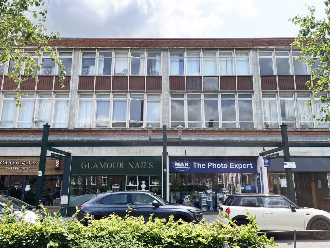 A front-on shot of a short and wide three storey building, with shops on the bottom floor and housing above. Cars are parked in front of the shops. The flats above feature almost full height, full width tall and narrow windows on their front. Trees partially frame the scene.