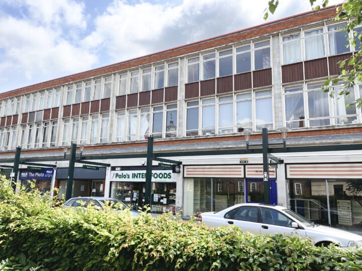 A two-storey building with large glass windows and a red roof. Ground floor shops include “Polos INTERFOODS” and “The Photo Shop.” Two white cars are parked in front, and green hedges line the street under a partly cloudy sky.