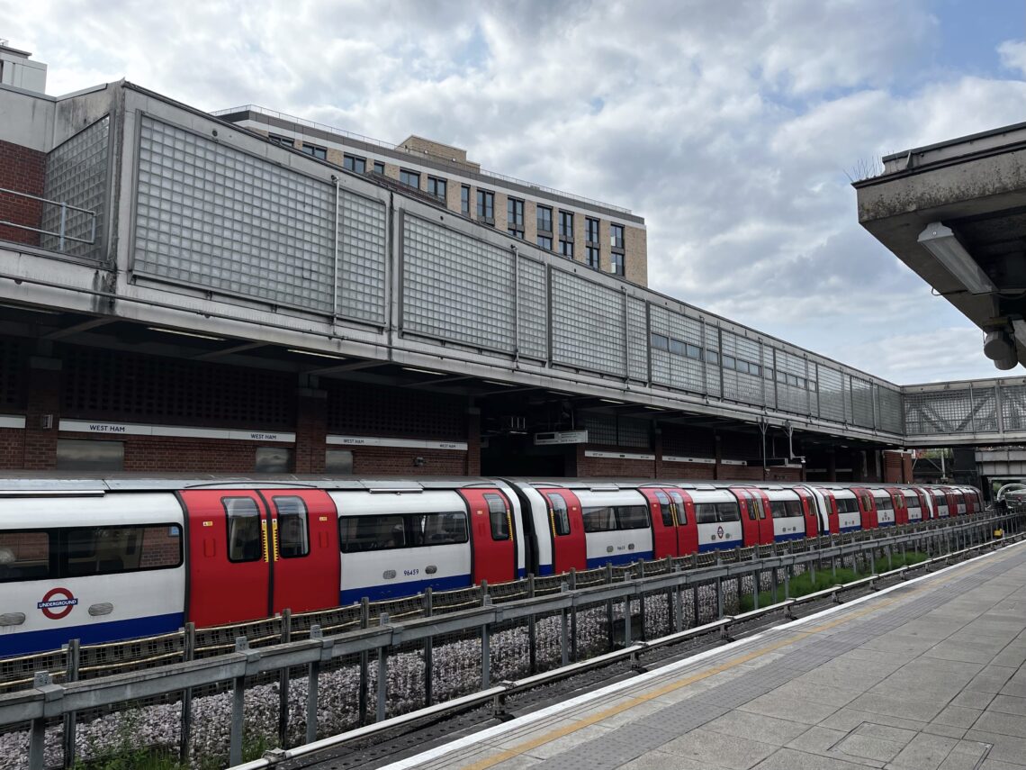 A red and white London Underground train is stopped at an outdoor station platform. Behind the train are brick and glass buildings. The sky is partly cloudy, and no people are visible on the platform or inside the train.