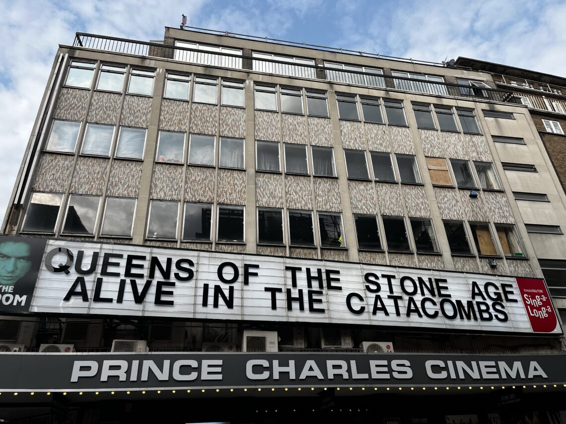 A marquee on the Prince Charles Cinema displays “QUEENS OF THE STONE AGE ALIVE IN THE CATACOMBS.” The cinema’s black awning is below, and the building above has patterned windows. The sky is partly cloudy.