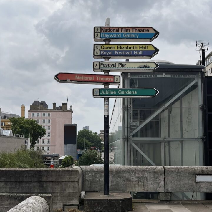 A signpost on a cloudy day points to nearby attractions including National Film Theatre, Hayward Gallery, Queen Elizabeth Hall, Royal Festival Hall, Festival Pier, National Theatre, and Jubilee Gardens, with buildings and trees in the background.