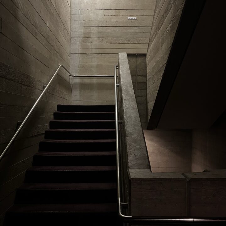 A dimly lit concrete stairwell with dark carpeted steps leads upward and turns left; stainless steel railings line both sides. The walls are textured concrete, and a small sign reading “STAIRS” is visible above the landing.