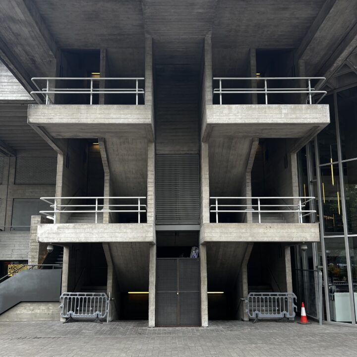 A brutalist concrete staircase with three steep flights leads to upper levels, framed by thick railings. Metal barriers and a traffic cone block the lower entrance. The building’s geometric, angular lines dominate the symmetrical design.