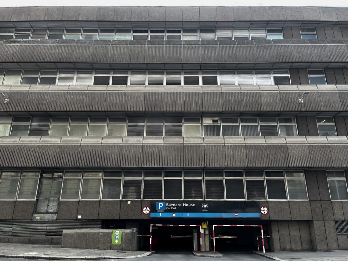 A building with 4 floors of continuous thin-framed aluminium windows, as well as vertical ribbed concrete between each floor. At the bottom, a series of metal horizontal vents and an entrance to an underground carpark.