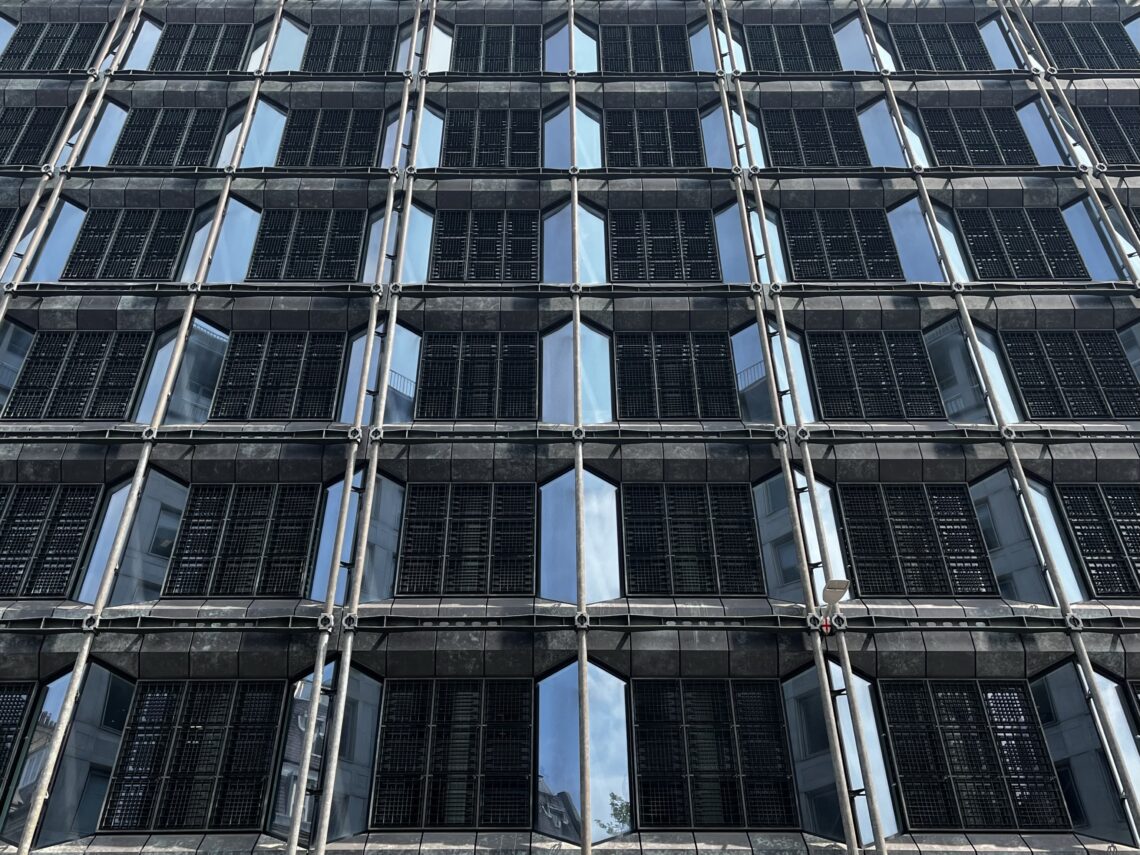 A modern glass building façade with a grid of repeating rectangular windows, each framed by metal and covered with dark, louvred shutters. The reflections of the sky and nearby structures are visible in the glass panels.
