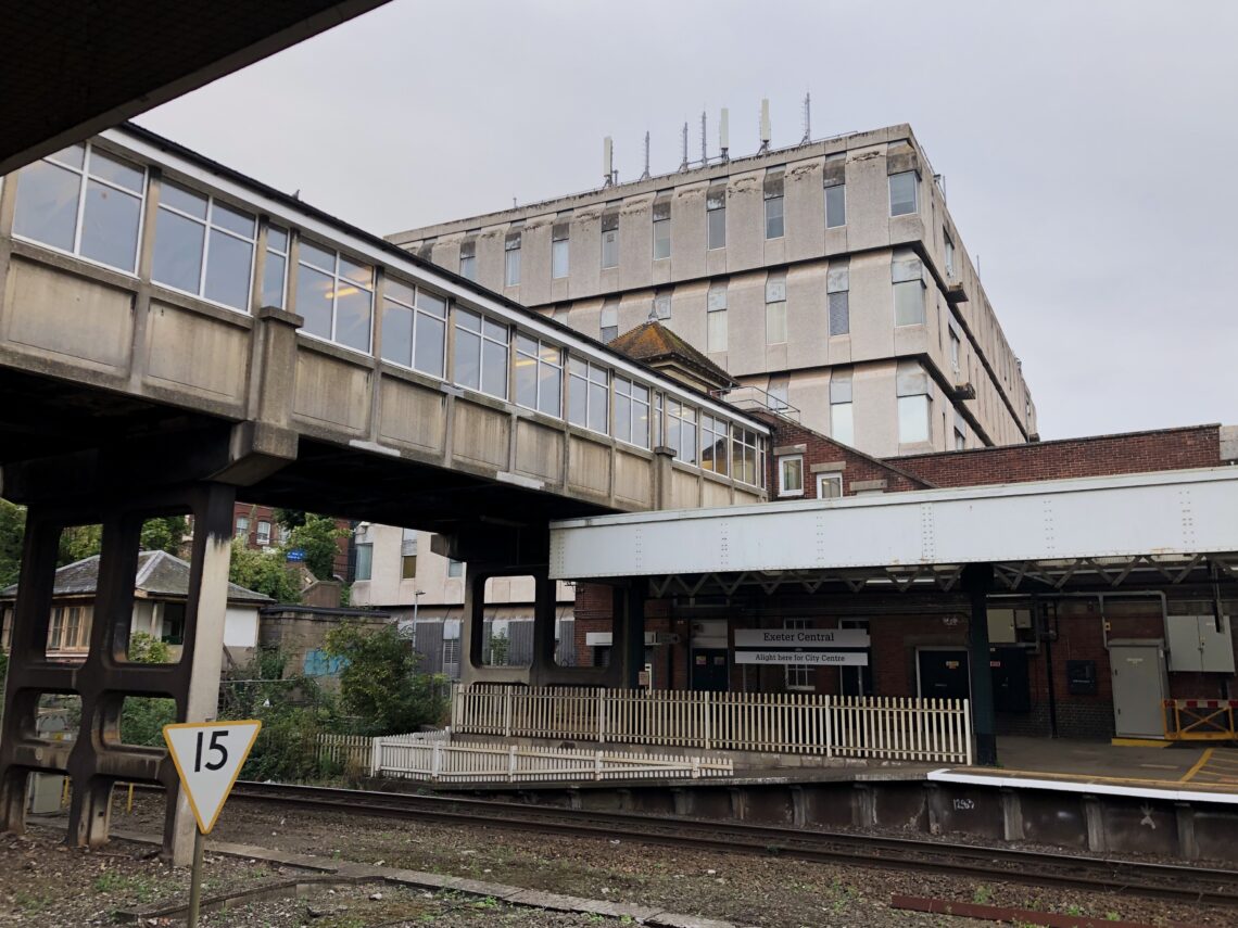 A railway station platform with a metal footbridge overhead, a large concrete building in the background, and a white sign displaying the number 15 near the tracks. The station appears empty and the sky is overcast.
