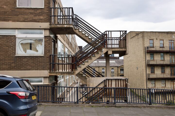 An exterior metal staircase with rusty railings is attached to a brick building. A broken window is visible on the left. A blue car is parked in front, and another tan brick building stands in the background under a cloudy sky.