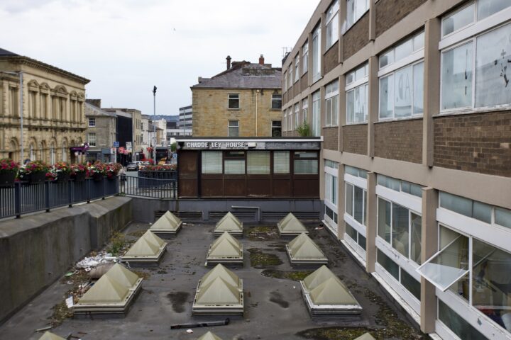 A rooftop area with pyramid-shaped skylights, some litter, and dirt. To the right is a brown and beige building with many windows, some open. In the background, there are older stone buildings, a street with people, and flower planters on a railing.