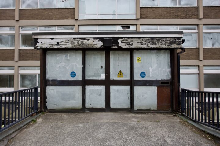 The image shows a dilapidated entrance to an old building. The doors are covered with frosted glass and graffiti, the paint on the canopy is peeling, and warning signs are visible. The walkway leading to the doors is bordered by black railings.