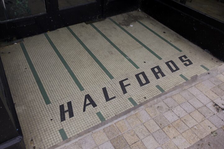Mosaic tile entrance with the word “HALFORDS” spelt in black tiles. The background is light-coloured with green vertical lines, leading to black-framed glass doors. Adjacent pavement consists of square beige and tan tiles.
