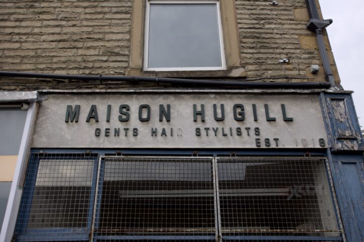 A weathered shop sign reads “MAISON HUGILL GENTS’ HAIR STYLISTS EST. 1918” above a metal-grilled window. The building exterior is stone with peeling blue trim and an upper window with a white frame, showing signs of age.