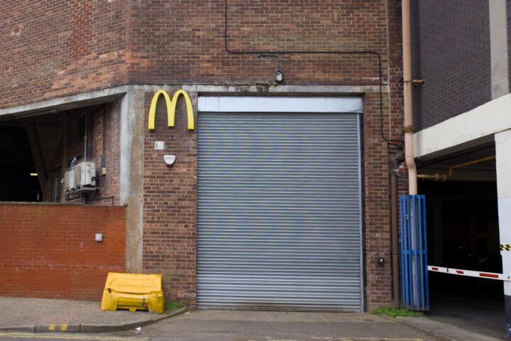 A large metal roller shutter is set into a brick building. Above the door is a yellow McDonald’s M logo, but there are no other restaurant signs. Nearby are a yellow bin, a red brick wall, and a blue gate leading to a car park.