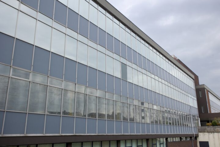A modern, multi-storey building with large glass windows reflecting the cloudy sky. The building has a rectangular shape and blue-tinted glass panels divided by vertical and horizontal frames, creating a grid-like appearance. The structure extends into the distance.