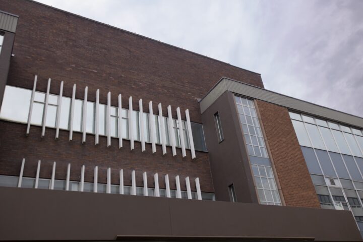 A modern brick building with large windows and vertical white metal bars on the façade. The sky is overcast, giving a muted tone to the scene. The architecture features both dark and light brown panels.