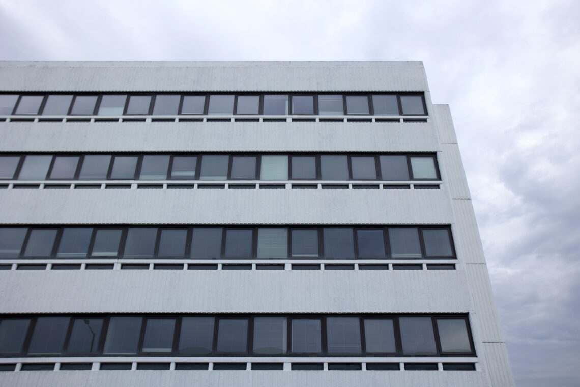A modern white building with flat, horizontal rows of rectangular, dark-framed windows. The facade is simple and geometric, stretching across several floors. The sky above is overcast, with grey clouds dominating the background.