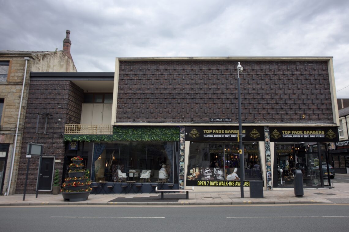 A modern barbershop with large glass windows and TOP FADE BARBERS signs above. Outdoor seating with tables and chairs is in front, along with a small decorative tree adorned with orange ornaments. The building has a brick façade and faces a quiet street.