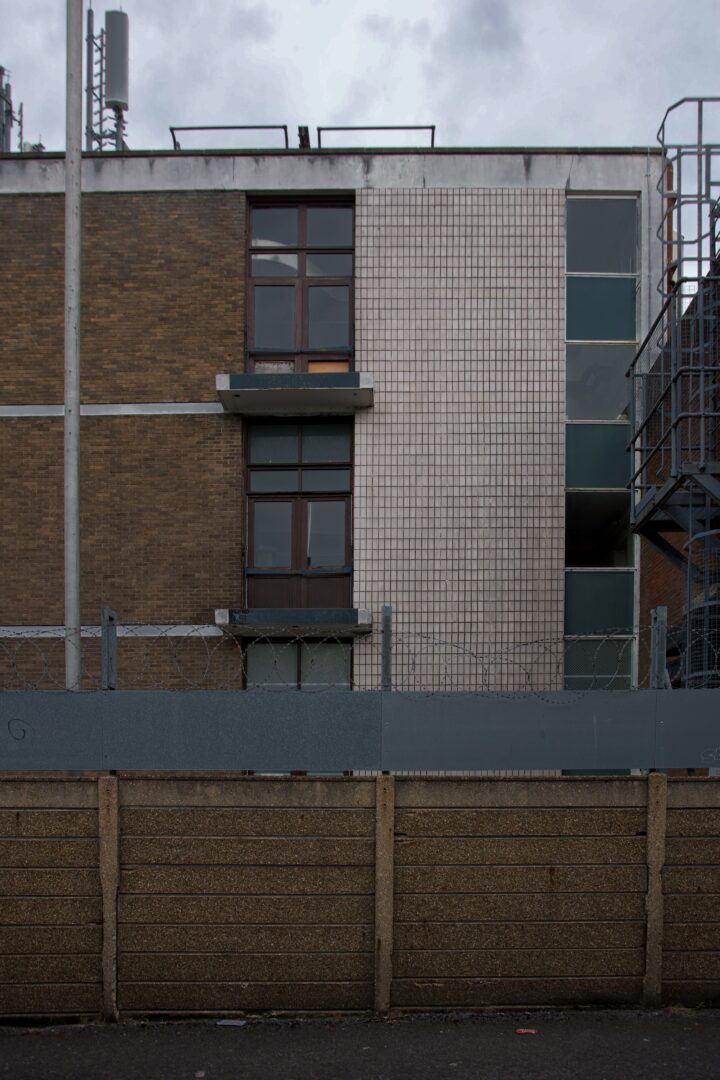 A three-storey building with brown brick and white tiled walls, tall windows, a metal staircase on the right, and aerials on the roof. The front is blocked by a high concrete and metal fence topped with barbed wire under a cloudy sky.