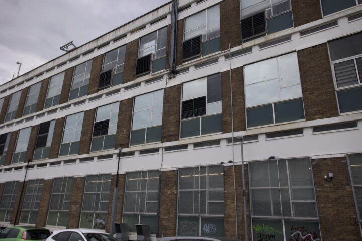 A three-storey building with brown brick and white trim, many large windows, some open or partially covered. The ground floor has graffiti on panels and several parked cars are visible in front. The sky is overcast.
