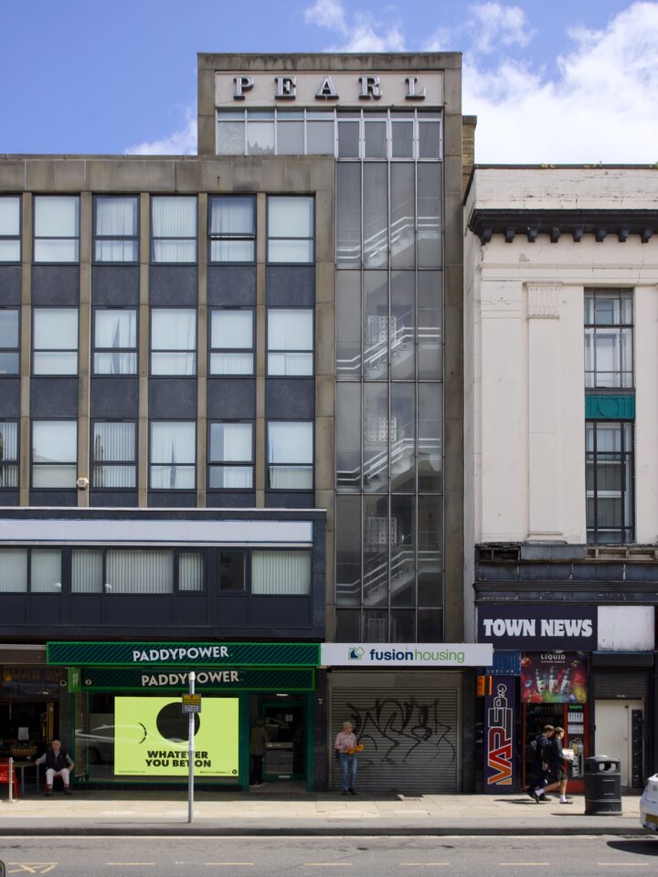 A city street scene showing a modern grey building labelled PEARL with glass-walled stairs, next to white and black older façades. Shops at street level include PADDYPOWER, fusion housing, and TOWN NEWS. A person stands outside, and someone sits at a table.