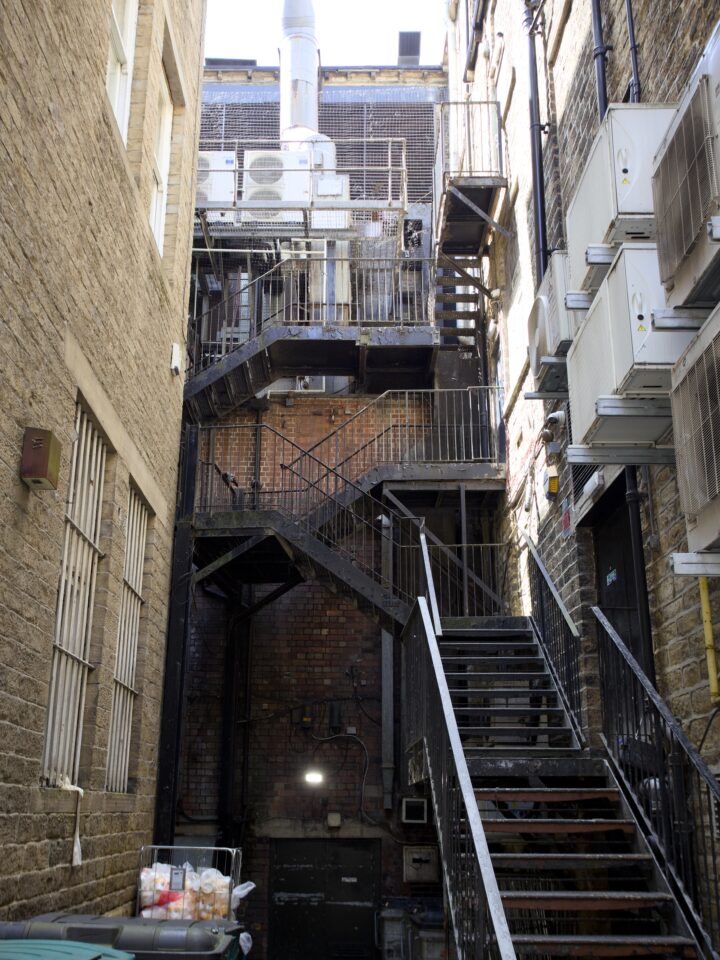 A narrow alleyway between stone buildings features a metal fire escape with zigzagging stairs and railings. Several air conditioning units are mounted on the right wall. Sunlight illuminates steps, and a rubbish bin with bags of bread sits in the left corner.