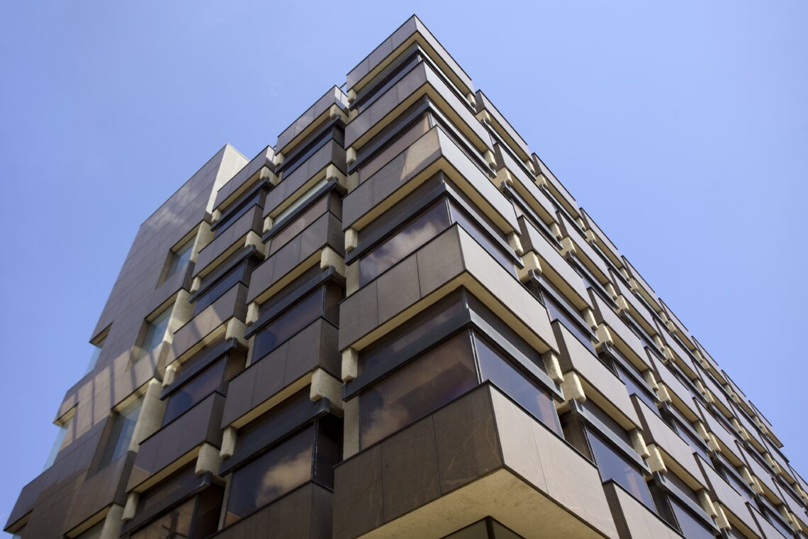 A modern building with brown-tinted glass windows and angular balconies, viewed from below against a clear blue sky. The structure features repeating geometric patterns and sharp lines, reflecting light and nearby surroundings.