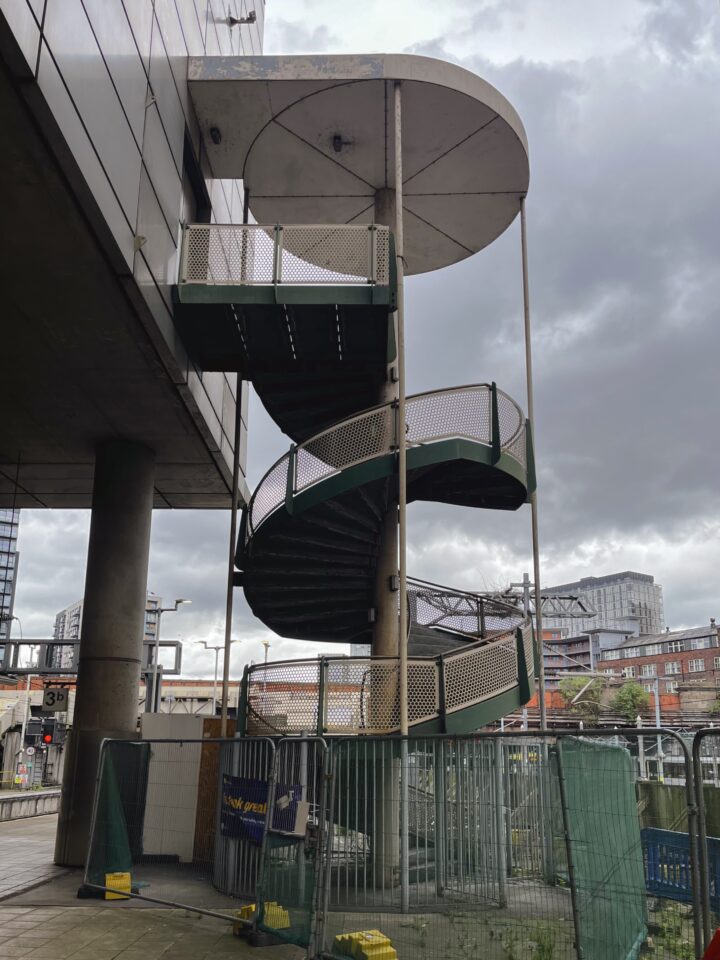 A green metal spiral staircase with mesh railings stands outdoors, sheltered partially by round roofs. It is attached to a modern building, next to a fence and construction barriers, with a cloudy sky and city buildings in the background.
