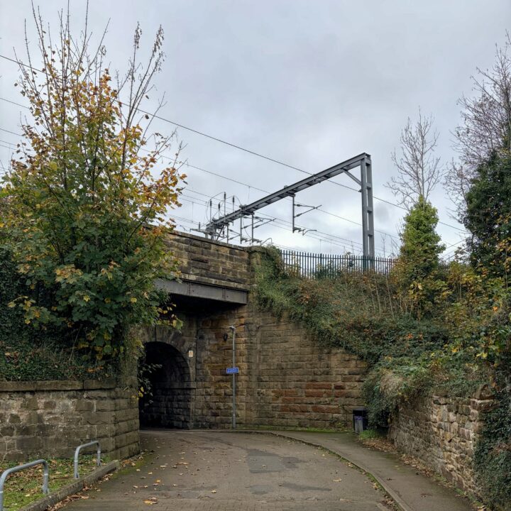 A narrow tarmac path curves under a stone railway bridge with overhead electric wires. Trees with autumn leaves and green bushes line the sides. A cloudy sky looms above. Metal railings and a blue sign are visible near the tunnel entrance.