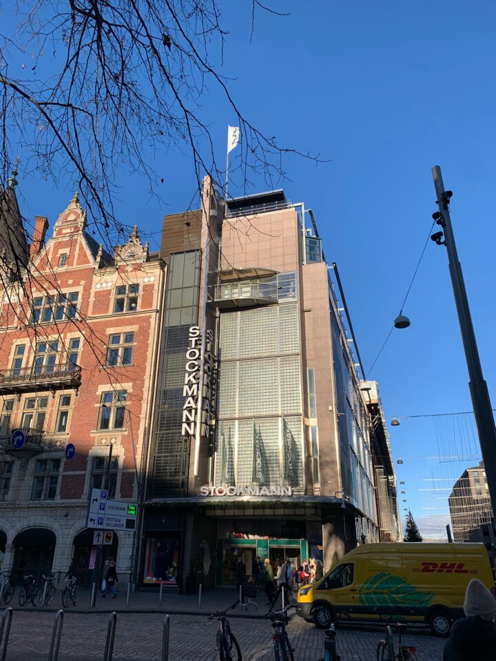 A modern multi-storey building with STOCKMANN signs stands beside an older red-brick building. A yellow DHL van is parked in front, and several bikes are lined up along the cobblestone street. The sky is clear and blue, with bare tree branches overhead.