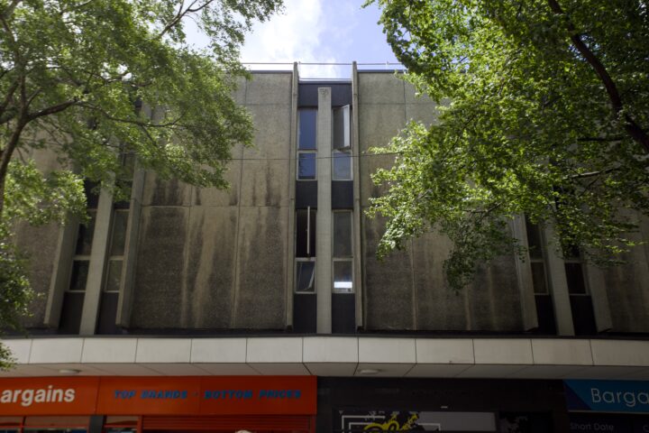 A grey, weathered concrete building with narrow vertical windows is partially shaded by leafy trees. At street level, there are red and blue shop signs, including one reading Bargains. The sky above is bright and partly cloudy.