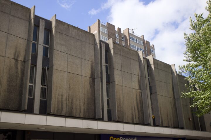 A Brutalist concrete building with tall, narrow vertical elements and dark stains beneath the windows. A modern block of flats is visible in the background, and a leafy tree is on the right under a partly cloudy blue sky.