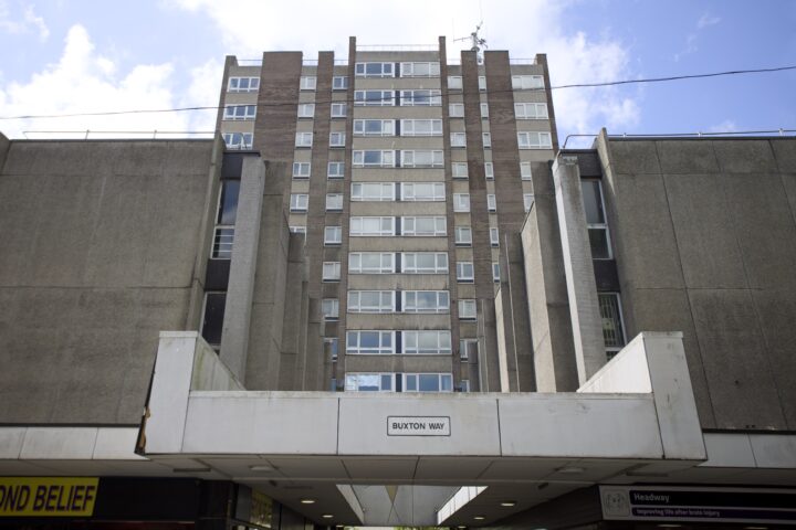 A tall, grey concrete block of flats with many windows stands behind a modern entrance labelled Buxton Way. The structure has a Brutalist architectural style, with boxy shapes and minimal decoration, beneath a partly cloudy blue sky.