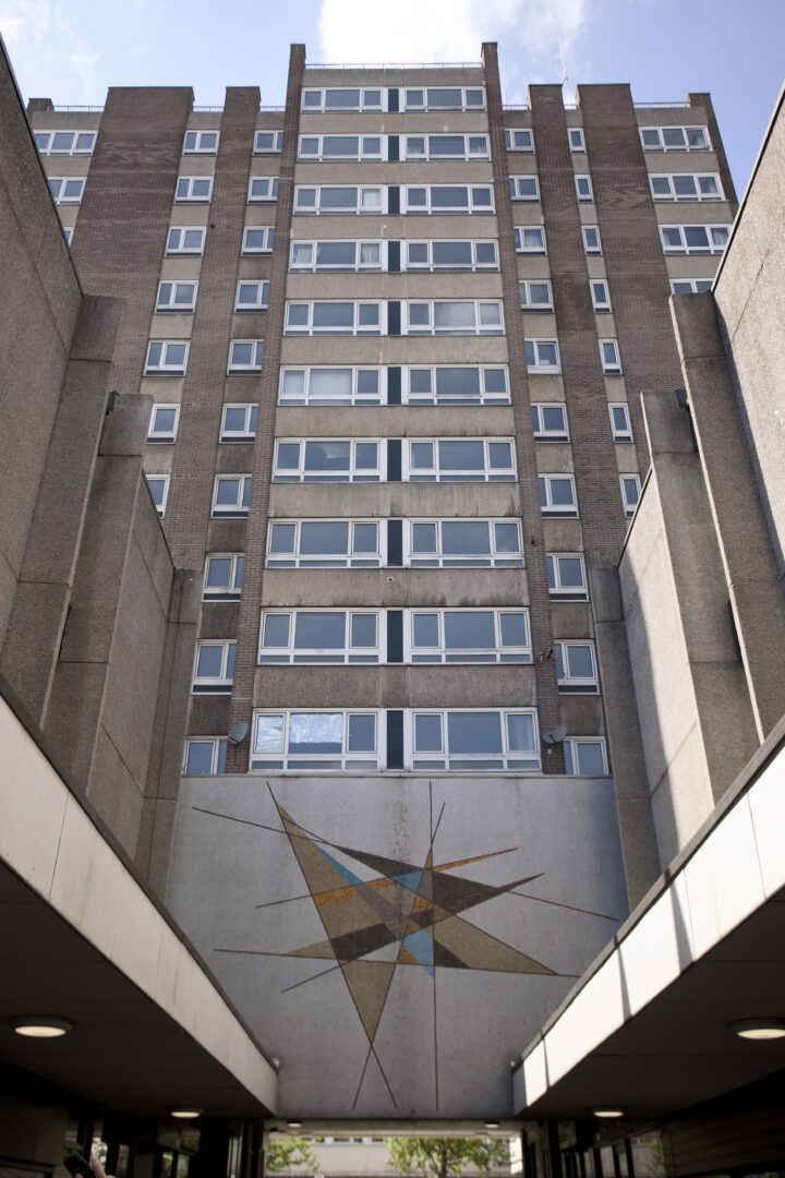 A tall, brown and grey block of flats with many rectangular windows rises into a blue sky. At the base, a geometric mosaic art piece decorates the wall, featuring intersecting triangles in shades of blue, yellow, brown, and cream.