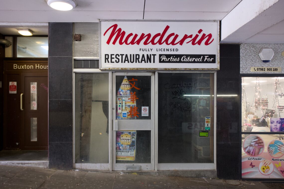 A closed restaurant with a sign reading Mandarin Restaurant, Fully Licensed, Parties Catered For above a glass door. Posters are on the door, and Chinese characters are visible on the window. A beauty salon and an entrance to Buxton House are nearby.