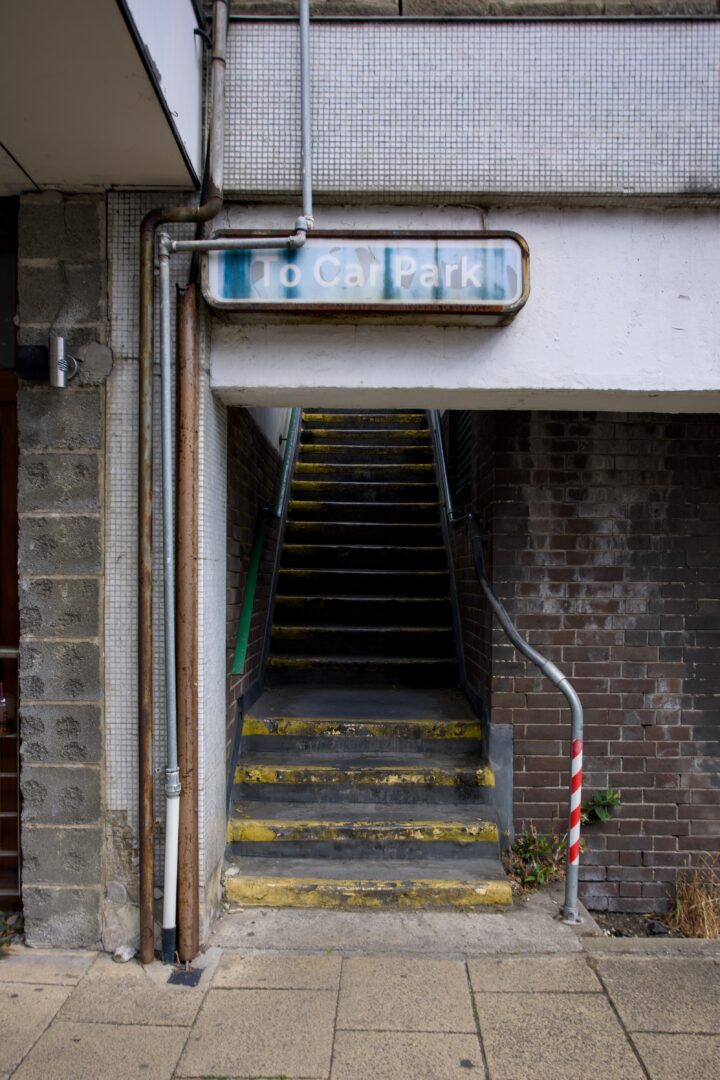 A faded sign reading “To Car Park” hangs above a narrow, worn staircase with yellow-striped steps. The stairs ascend between old brick and concrete walls. A metal handrail lines the right side, next to a red and white striped pole on the pavement.