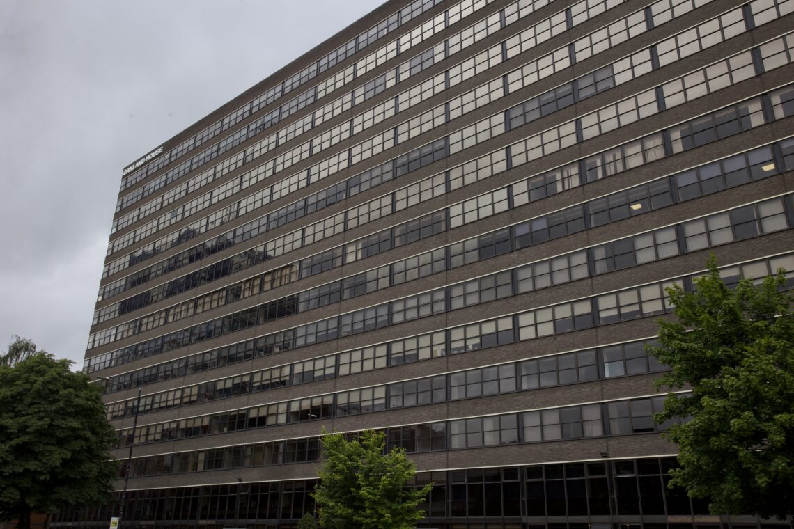 A large, multi-storey office building with many rows of windows and a brick façade, viewed from a low angle. Trees with green leaves are in the foreground, and the sky above is overcast and grey.
