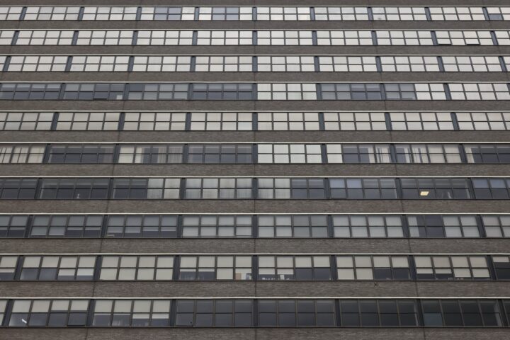 A close-up view of a modern building’s façade, showing multiple rows of rectangular windows set in dark frames. The walls between the windows are made of brown brick, creating a striped pattern with the alternating glass panes.