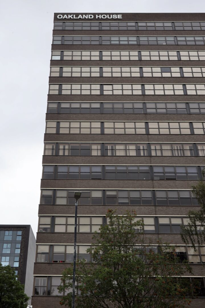 A tall, multi-storey office building with rows of large windows. The top of the building has the sign OAKLAND HOUSE. Trees and a streetlamp are visible in the foreground, and the sky behind is overcast.