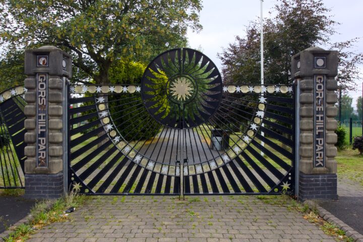 Black metal gates with a sunburst design and star accents mark the entrance to Gorse Hill Park. “Gorse Hill Park” is displayed vertically on stone pillars on each side. Trees and a paved path are visible beyond the gates.