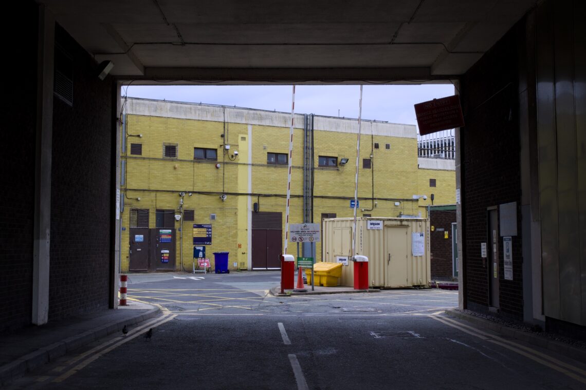 A view through a dark underpass opens onto a fenced industrial area with yellow-brick buildings, various warning signs, bollards, a yellow grit bin, and a beige shipping container. The scene is empty, with overcast sky and muted colours.