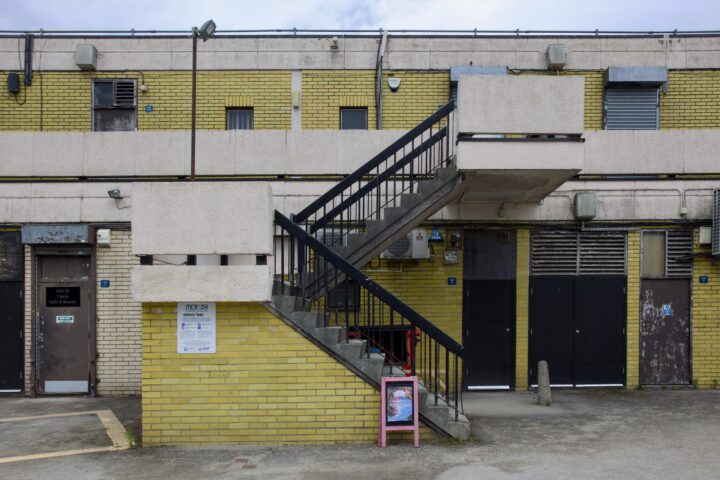 A yellow-brick building with a concrete and metal staircase leading to the upper floor. Black doors and vents line the ground floor, and a small framed sign sits near the stairs. The scene looks urban and slightly worn, under a cloudy sky.