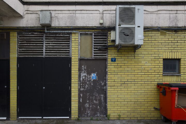 A yellow brick wall with a rusty metal door, a blue sign, barred vents, and a large white air conditioning unit mounted above. To the right, a red skip sits beneath a small barred window. The area is unused, with some peeling paint and overgrown weeds.