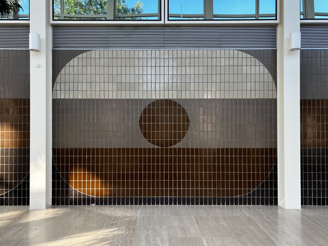 A tiled wall features a large geometric design: a big white semicircle fills most of the space, with a centred brown circle inside it. The tiles are arranged in a grid, and sunlight streams in from upper windows above the artwork.