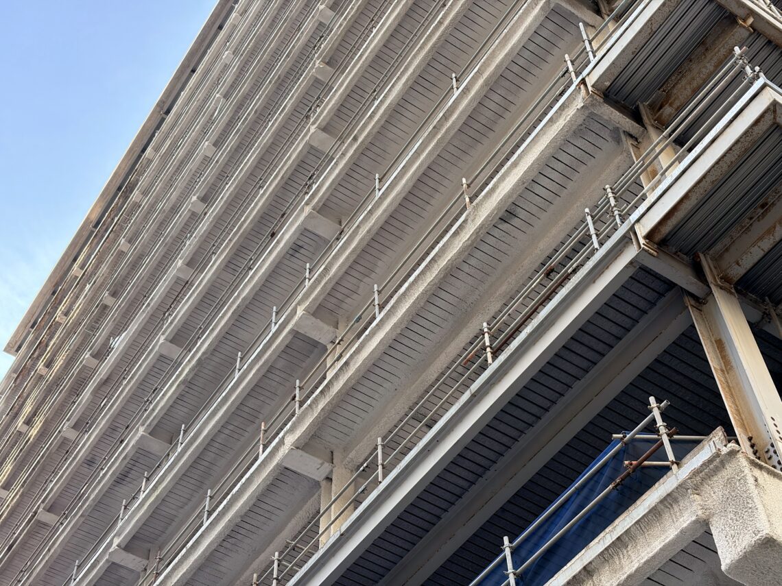 Close-up view of the exterior of a multi-storey concrete car park, showing rows of repetitive horizontal balconies with metal railings. The image is taken from a low angle, looking up, highlighting the structure’s geometric lines and weathered surfaces.