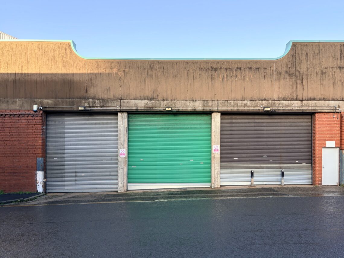 A building with three large metal roller doors—grey, green, and brown—set in a weathered façade. Brick walls frame the sides, and a wet street is in front. The sky is clear, and two small “No Parking” signs are visible on either side of the green door.
