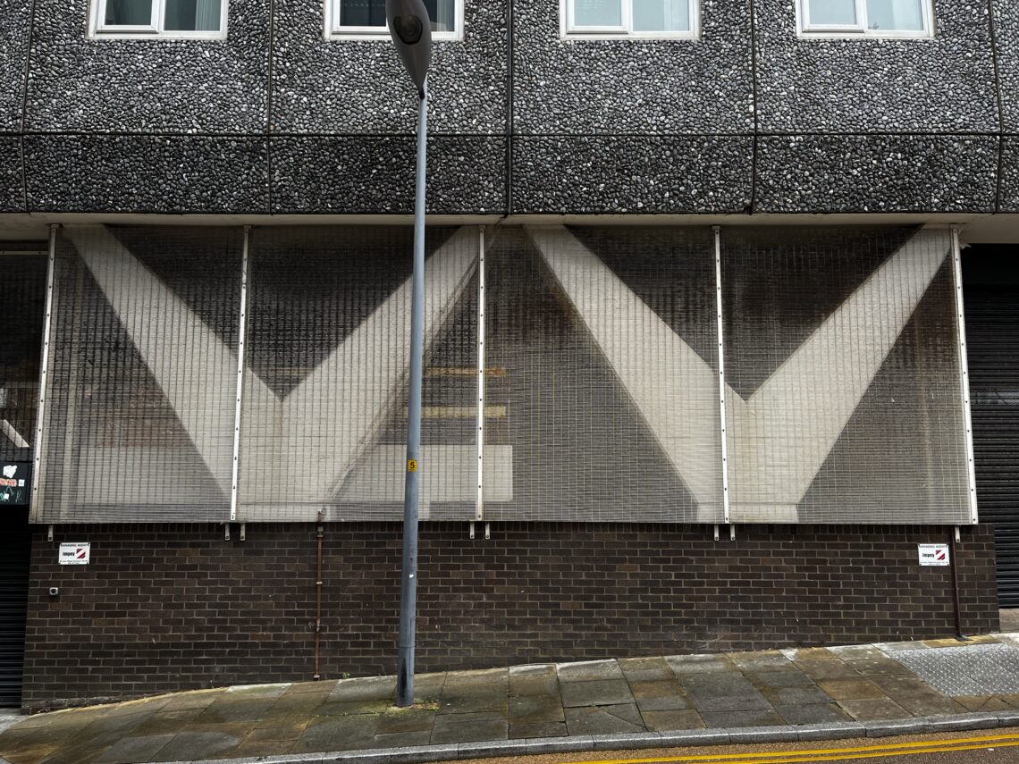 A textured building façade features three large mesh panels with bold white “V” shapes. A streetlamp stands in front, casting a shadow. The pavement below is wet, and two small no parking signs are visible on the dark brick wall.